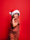 Adorable petite fille en vêtements décontractés et chapeau de Père Noël boudant lèvres et toucher le visage tout en se tenant debout sur fond rouge et en regardant la caméra — Photo de stock