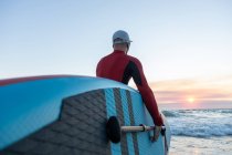 Visão traseira do surfista masculino irreconhecível em roupa de mergulho e chapéu carregando prancha de remo e entrando na água para surfar na costa — Fotografia de Stock