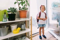 Lächelndes Schulkind mit Rucksack und Schleife am Haar, das daheim zwischen Glastür und Topfpflanzen in die Kamera blickt — Stockfoto