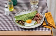Домашній сніданок шпинатних млинців з беконом, яйцем та грибами, подається на білій тарілці з сіллю та масляним шейкером на картатому скатертині . — стокове фото