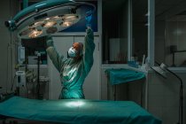 Молодая женщина ветеринар в хирургической форме и стерильной маске глядя вверх, регулируя лампу над медицинским текстилем в клинике — стоковое фото
