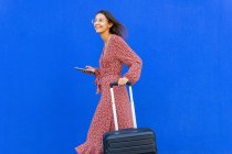 Vista lateral de la mujer positiva en vestido largo rojo caminando con el equipaje mientras navega en el teléfono inteligente en la calle contra la pared azul en el día - foto de stock