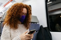 Mulher atenta com cabelos encaracolados em pano máscara facial navegar na internet no celular durante a viagem de trem — Fotografia de Stock