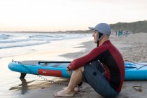 Side view of thoughtful male surfer in wetsuit and hat sitting looking away with SUP board while preparing to surf on seashore — Stock Photo