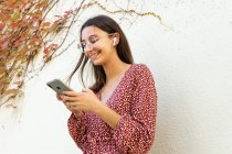 Весела жінка в повсякденному одязі, що з'єднує бездротові навушники зі смартфоном проти світлої стіни вдень — стокове фото
