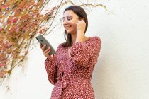 Femme gaie en vêtements décontractés connectant des écouteurs sans fil avec smartphone contre un mur de lumière pendant la journée — Photo de stock