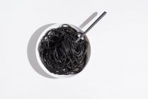 Vue du dessus de la fourchette en acier inoxydable dans un bol plein de spaghettis noirs avec de l'encre de seiche sur une table blanche en studio — Photo de stock