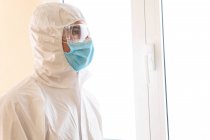 Adult male medic in personal protective equipment with glasses and sterile mask looking forward against window in hospital — Stock Photo