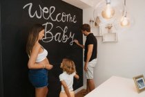 Male writing Welcome Baby inscription on blackboard against expectant female beloved and daughter in house — Stock Photo