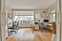 Intérieur de la pièce contemporaine avec ordinateur de bureau sur table contre fauteuil et chaises sur tapis dans la maison lumineuse — Photo de stock