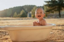 Vue latérale de l'enfant tout-petit heureux avec jouet assis regardant loin dans le bain en plastique tout en jouant avec l'eau dans la campagne — Photo de stock