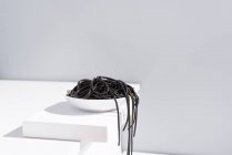 Estudio minimalista con espaguetis de tinta de calamar negro cayendo desde un tazón de cerámica completo sobre una mesa blanca - foto de stock