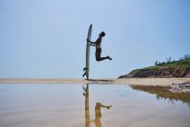 Vista laterale di felice surfista afro-americana con longboard surf saltando sopra la costa sabbiosa sotto il cielo blu — Foto stock