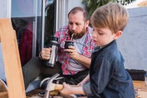 Hipster dad pouring herbal tea from thermos and drinking in calabash gourd against son with hammer working with wood — Stock Photo