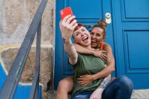 Cheerful homosexual women with tattoos taking self portrait on cellphone against entrance door in town — Stock Photo