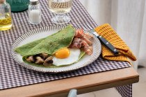 Pequeno-almoço caseiro de panquecas de espinafre com bacon, ovo e cogumelos servido em uma placa branca com um sal e óleo agitador em uma toalha de mesa quadriculada — Fotografia de Stock