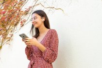 Cheerful female in casual clothes connecting wireless earphones with smartphone against light wall in daytime — Stock Photo