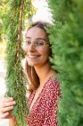 Joyeux jeune femme avec les cheveux bruns dans les lunettes debout parmi les branches vertes et regardant la caméra en plein jour — Photo de stock
