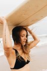 Nachdenkliche junge Sportlerin in Badebekleidung mit fliegendem Haar, die ein Surfbrett auf dem Kopf trägt und auf die Meeresküste blickt — Stockfoto
