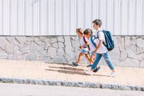 Vista lateral del colegial con mochila hablando con amigas mientras pasea sobre pavimento de baldosas contra pared de piedra a la luz del sol - foto de stock