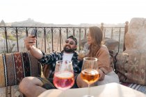 Positives junges stilvolles Paar, das auf der Couch sitzt und Cocktailgläser klimpert, während es auf der Terrasse in Kappadokien, Türkei, ein Selbstporträt auf dem Handy macht — Stockfoto