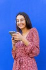 Mulher sorridente em vestido e óculos de pé perto da parede azul e usando smartphone durante o dia — Fotografia de Stock