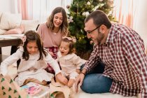 Здивовані дівчата з веселими батьками відкривають коробку з подарунками на підлозі під час святкування різдвяного дня у кімнаті. — стокове фото