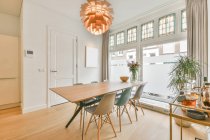 Сучасний інтер'єр їдальні з дерев'яним столом і пластиковими стільцями під творчою люстрою в просторій новій квартирі — стокове фото