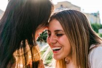 Cheerful teen covering mouth while whispering in ear of best female friend with closed eyes in sunlight — Stock Photo