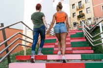 Vista posterior de mujeres homosexuales de moda anónimas cogidas de la mano mientras suben por la escalera contra casas urbanas - foto de stock
