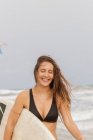 Young cheerful sportswoman with flying hair and surfboard in ocean with foam under cloudy sky — Stock Photo
