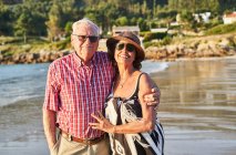 Sorridente coppia anziana a piedi nudi in occhiali da sole in piedi sulla spiaggia di sabbia bagnata e godersi la giornata di sole — Foto stock
