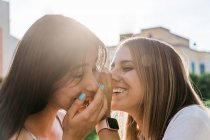 Adolescente alegre cobrindo a boca enquanto sussurra no ouvido do melhor amigo do sexo feminino com os olhos fechados na luz solar — Fotografia de Stock