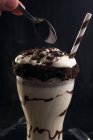 Crop pessoa anônima com colher sobre o vidro do delicioso milkshake decorado com biscoitos de chocolate esmagado no fundo preto — Fotografia de Stock