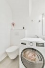 Интерьер светлой ванной комнаты с современной стиральной машиной расположен возле раковины и зеркала в ванной комнате с душевой кабиной — стоковое фото