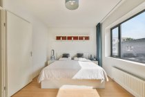 Cama com almofadas sob prateleira e lâmpada acima parquet na casa moderna com janela e radiador no dia ensolarado — Fotografia de Stock