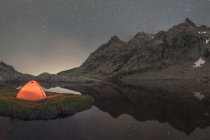 Vista panorâmica da tenda na costa do lago contra a montanha nevada sob o céu nublado à noite — Fotografia de Stock