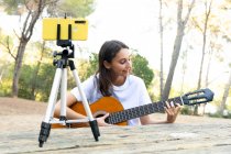 Весела жінка-підліток блогер грає на акустичній гітарі під час запису відео на мобільний телефон на тринозі в парку — стокове фото