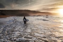 Vista posteriore di una giovane donna con tavola da surf che entra in mare durante il tramonto sulla spiaggia delle Asturie, Spagna — Foto stock