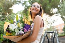 From below content young female in eyeglasses looking away standing with blossoming flower bouquet on urban stairs — Stock Photo