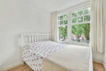 Interior minimalista de dormitorio con cama cómoda contra ventanas y paredes de luz durante el día - foto de stock