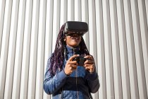 Задоволена молода афроамериканська жінка в VR-хедсеті з використанням контролера під час розваги і гри в віртуальну гру проти сірої смугастої стіни — стокове фото