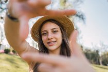 Усміхнена жінка-підліток з брекетами, що демонструють фотографічний жест, дивлячись на камеру вдень на розмитому фоні — стокове фото