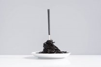 Нержавіюча виделка в повному посуді з чорного спагетті з каракатицьким чорнилом на білому столі в студії на сірому фоні — стокове фото