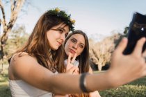 Female teens demonstrating victory gesture while taking self portrait on cellphone in sunny park on blurred background — Stock Photo
