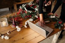 Обрізані невпізнавані жінки-флористи, що стоять за столом і використовують ноутбук, роблячи різдвяні прикраси в стильній квітковій студії — стокове фото