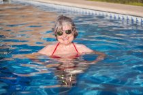 Cheerful elderly female with gray hair swimming in pool and brightly smiling at camera with sunglasses — Stock Photo