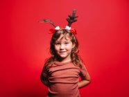 Gioiosa bambina in abiti casual e fascia festiva cervo guardando la fotocamera durante la celebrazione di Natale sullo sfondo rosso — Foto stock