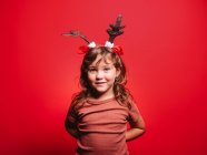 Alegre niña en ropa casual y diadema de ciervo festivo mirando a la cámara durante la celebración de Navidad contra el fondo rojo - foto de stock