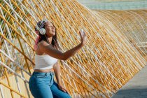 Jovem mulher étnica positiva com celular tirando uma selfie enquanto ouve música de fones de ouvido sem fio enquanto ri na cidade — Fotografia de Stock